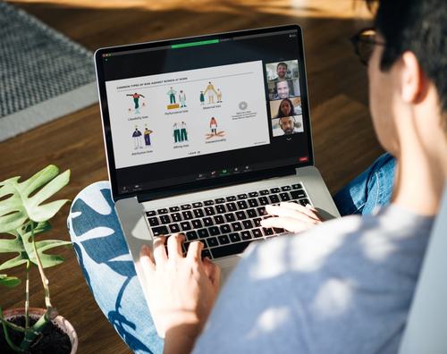 A person sits cross-legged on the floor using a laptop during a video call, viewing a presentation on "Common Types of Bias Against Women at Work" with illustrated figures, while four participants are visible in a sidebar.