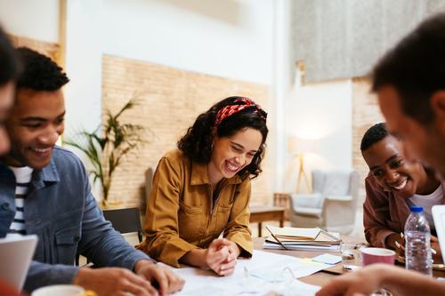 A diverse group of young people laugh and collaborate around a table covered with papers and notes in a bright, modern space. A smiling woman in a yellow jacket wearing a red headband is the focal point.