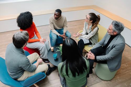 A diverse group of six people sit in a circle on colorful chairs around a small round table, engaged in conversation. The setting appears to be a modern office or collaborative workspace with wooden floors.