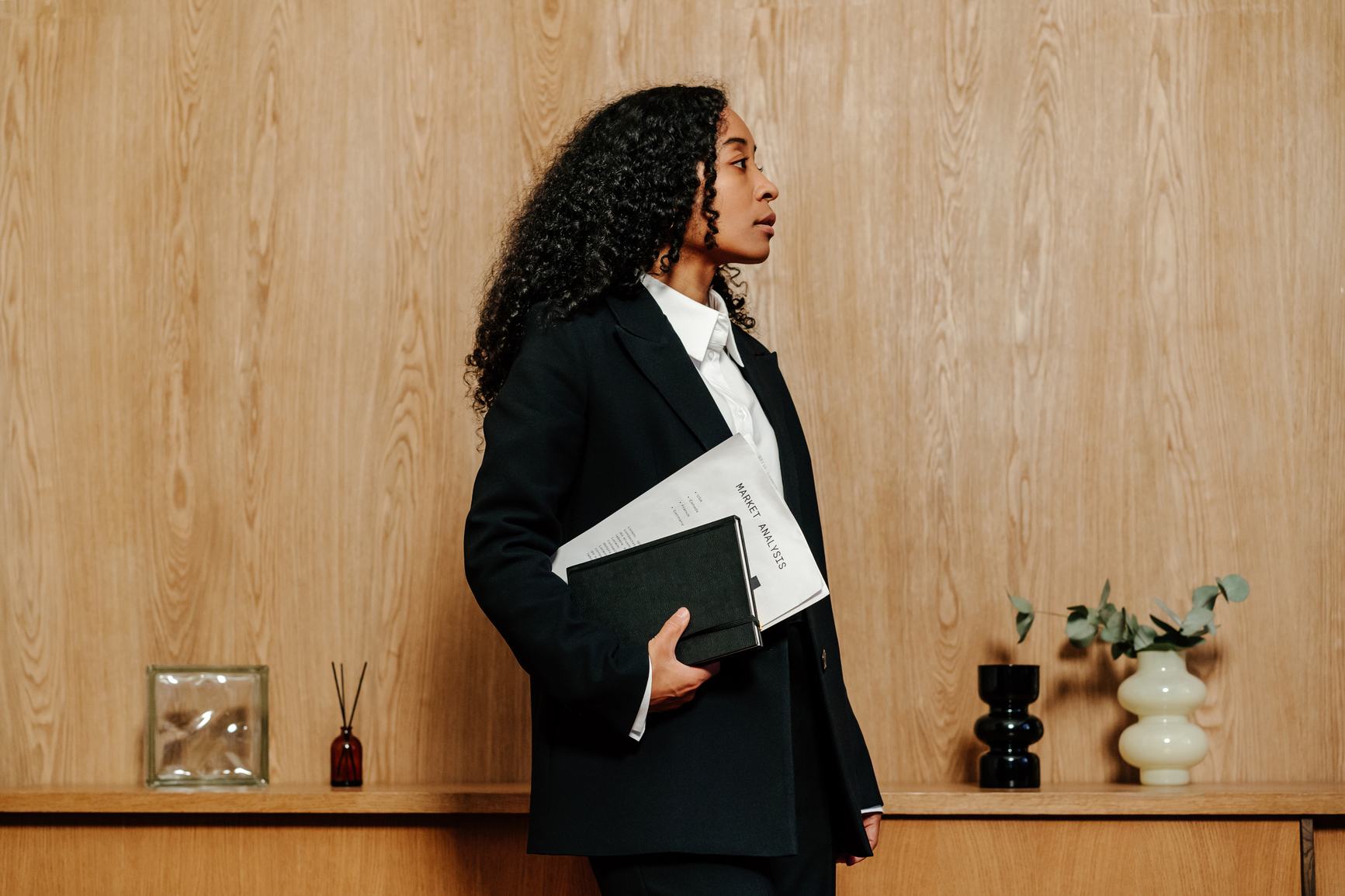 A professional woman with curly hair, wearing a black blazer, holds a notebook and market analysis documents. She stands in profile against a wooden wall, with decorative vases and a reed diffuser on a shelf behind her.