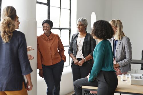 A diverse group of five professional women engaged in a friendly conversation in a modern, bright office setting, smiling and networking in a relaxed, collaborative atmosphere.