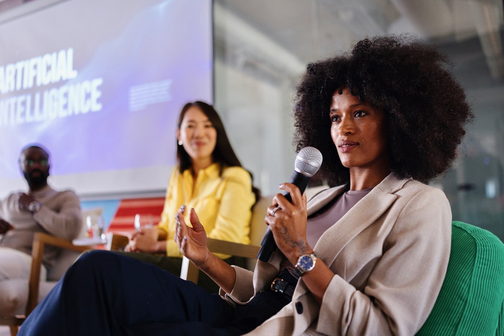 A woman with an afro speaks into a microphone at an Artificial Intelligence panel discussion, with two other panelists seated behind her and an AI-themed presentation slide visible in the background.
