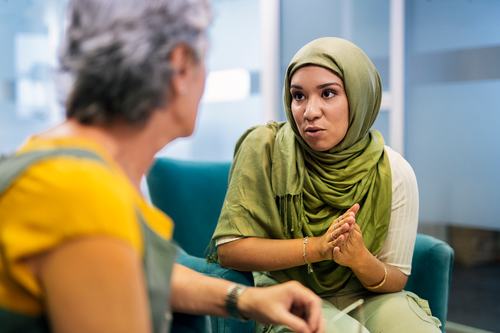 A young woman wearing a green hijab speaks earnestly to an older woman in a teal chair, hands clasped, in what appears to be a professional or counseling setting.