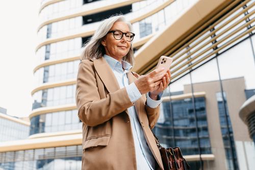 A well-dressed older woman with gray hair and glasses smiles while using her smartphone outdoors, wearing a beige blazer and carrying a brown bag, with a modern glass building in the background.