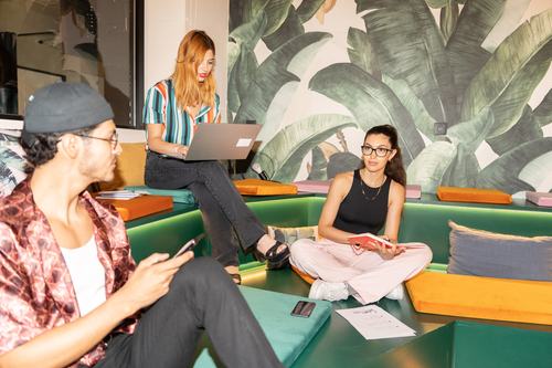 Three young people in a colorful lounge with a tropical leaf mural. One uses a laptop, one holds a red book, and one checks a phone, all seated on green cushioned benches with vibrant orange and yellow accents.