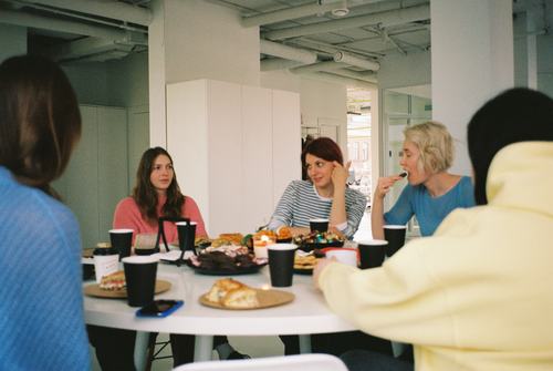 A group of women gathered around a white table in a bright office space, enjoying snacks and coffee. They appear to be chatting and relaxed, with food and black paper cups spread across the table.