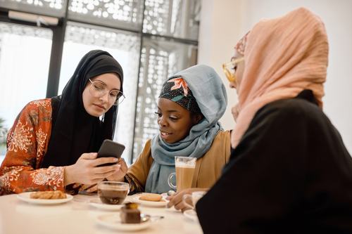Three women wearing hijabs gather at a café table with drinks and snacks, looking at a smartphone together and smiling.