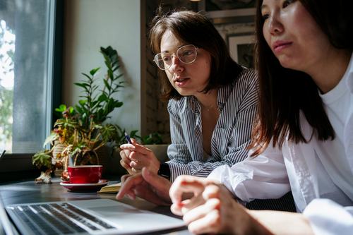 Two women collaborate at a cafe table with a laptop. One wears glasses and a striped shirt, holding a pen while speaking. A red coffee cup and green plants are visible near a sunlit window.