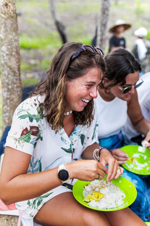 A smiling woman with sunglasses on her head eats rice from a green plate outdoors. She wears a floral top and a smartwatch. Others sit nearby, enjoying a casual outdoor meal among trees.