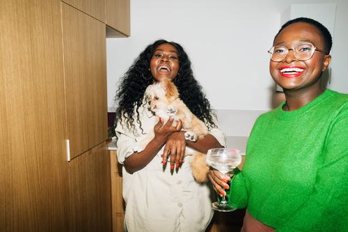 Two joyful women laugh together indoors; one holds a fluffy puppy while the other, wearing a green sweater and glasses, holds a cocktail glass. Both have red nails and bright smiles in a modern interior.