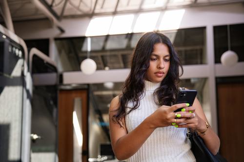 A young woman with long dark hair, wearing a white sleeveless turtleneck, looks at her smartphone. She has bright green nails, a gold bracelet, and carries a black bag, standing in a modern, well-lit indoor space.