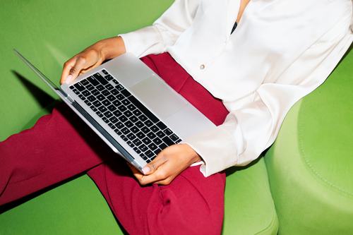 A person in a white blouse and red pants sits on a green chair, holding a silver laptop open on their lap against a vibrant green background.