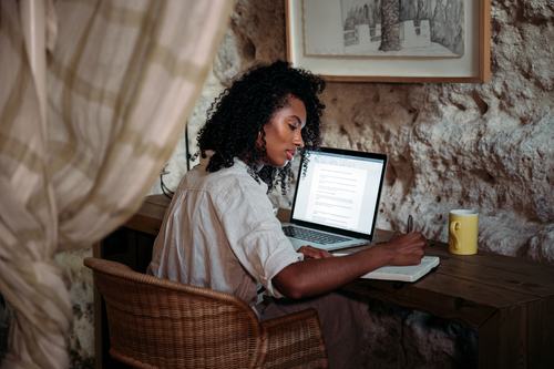 A woman with curly hair sits at a rustic wooden desk, writing in a notebook while referencing a laptop. She's in a cozy stone-walled room with a framed artwork, curtain, and yellow mug nearby.