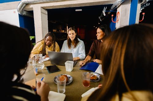 A group of women gathered around a table in a modern workspace, collaborating over a laptop. Drinks, snacks, a tablet, and notebooks are on the table, suggesting a casual business meeting or brainstorming session.