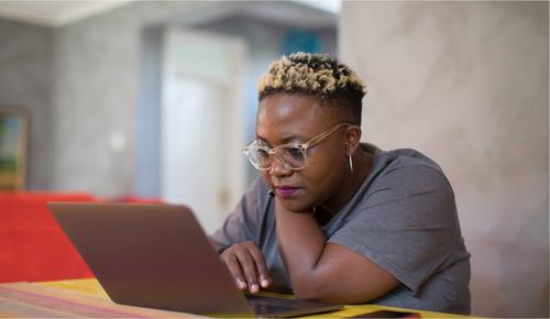 A woman with blonde-tipped natural hair and clear-framed glasses leans on a table, focused on her laptop screen. She wears a gray t-shirt and hoop earrings, working in a bright, modern indoor setting.