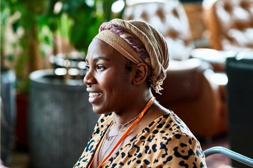 A smiling woman wearing a beige head wrap and leopard-print top with an orange lanyard, seated in a stylish indoor space with lush green plants and leather chairs in the background.