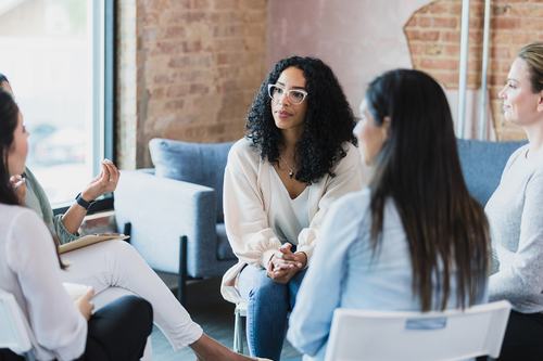 A group of women sit in a circle, engaged in discussion in a cozy, modern space with exposed brick walls. A woman with curly hair and white glasses sits attentively, hands clasped, as others listen and speak.