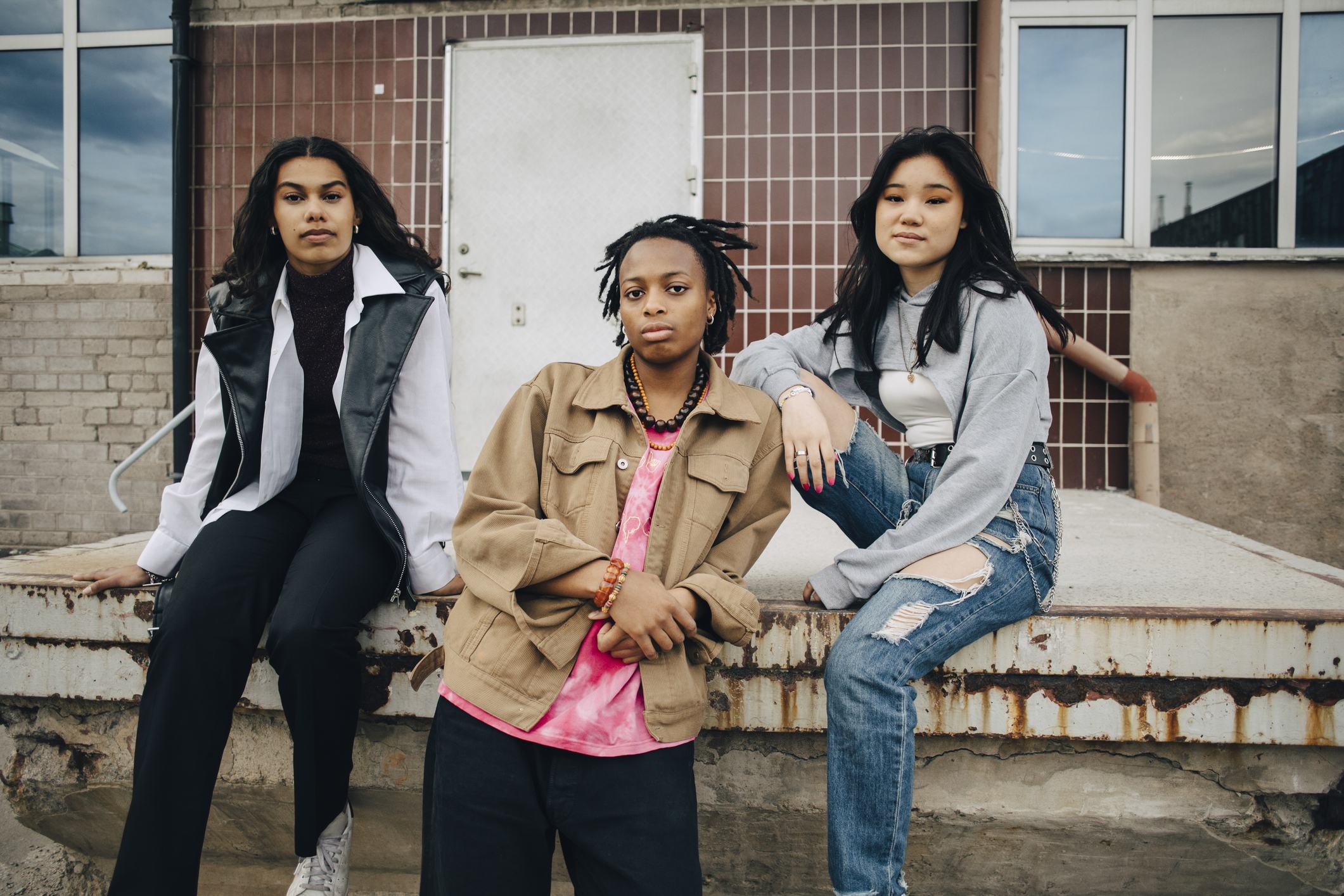 Three young women pose confidently in front of an urban building. They wear casual streetwear—a leather vest, a tan jacket over a pink tee, and a gray sweatshirt with ripped jeans—exuding a cool, edgy vibe.