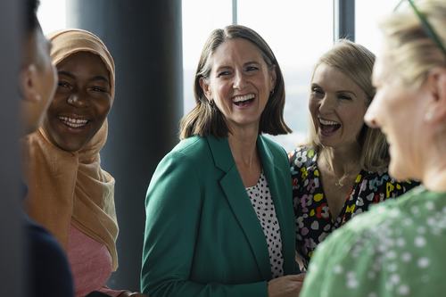A diverse group of women laughing and smiling together in a bright indoor setting, conveying warmth and camaraderie. They wear colorful outfits, including a green blazer and a hijab, suggesting a friendly professional or social gathering.