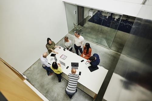 Aerial view of a diverse group of seven professionals gathered around a white conference table with laptops and tablets, collaborating in a modern office space with glass walls.