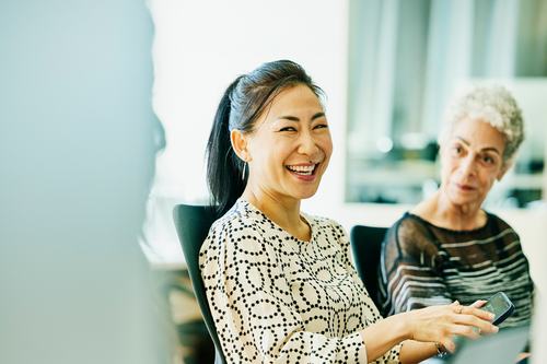 A smiling woman with dark hair in a patterned blouse sits in an office setting, holding a phone. An older woman with gray hair sits beside her. Both appear engaged in a professional meeting.