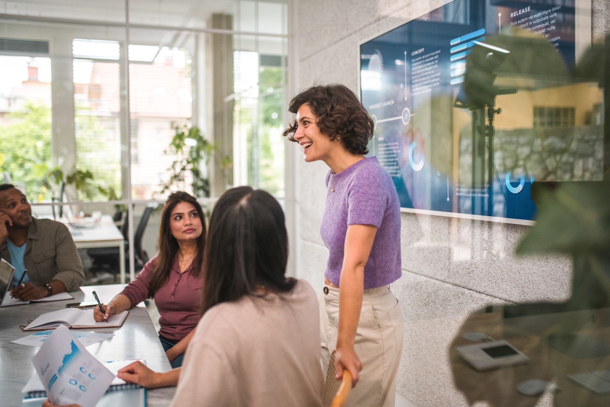 A smiling woman in a purple top leads a business presentation in a bright office, standing before a digital screen while colleagues seated at a table listen and take notes.