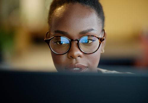 A close-up of a focused young woman wearing tortoiseshell glasses, intently looking at a screen. Blue light reflects off her lenses, highlighting her concentration against a softly blurred background.