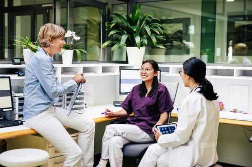 Three healthcare professionals—a woman in a blue shirt, a nurse in purple scrubs, and a doctor in a white coat holding a tablet with X-rays—share a cheerful conversation at a bright, plant-filled medical workstation.