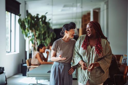 Two women laugh together in a bright, modern office. One wears a gray top and trousers; the other wears a green blouse with red braids. A colleague works in the background near large windows and lush plants.