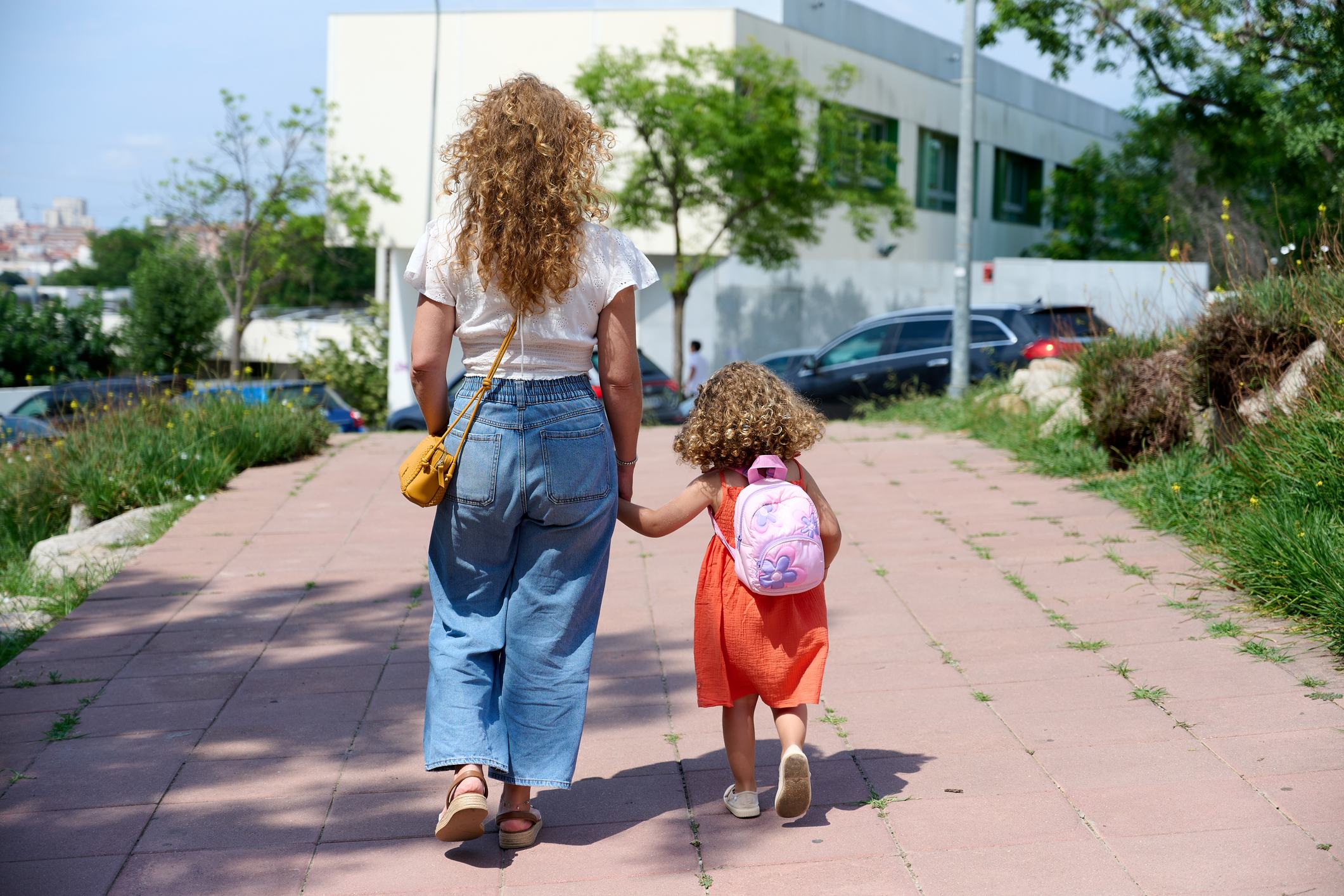 A woman with curly hair in jeans and a white top walks hand-in-hand with a young girl in an orange dress carrying a pink backpack, both seen from behind on a brick path near a building.