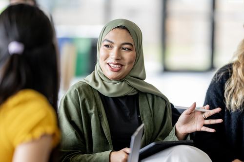 A young woman wearing a green hijab and jacket smiles warmly while gesturing during a conversation with others. She holds a tablet, suggesting a collaborative or educational setting.