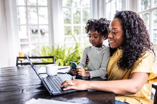 A smiling woman in a yellow top works on a laptop at a wooden table while a young child sits beside her holding a smartphone. A coffee cup and papers are nearby, with bright windows in the background.
