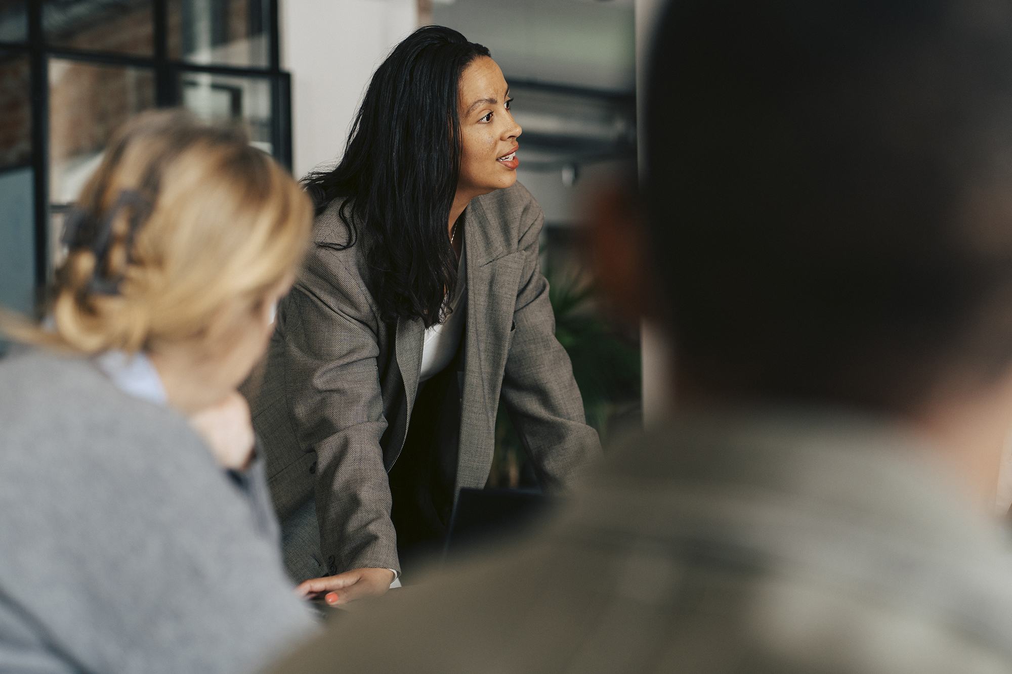 A businesswoman in a gray blazer leans forward, speaking confidently to colleagues in a modern office setting. Two people are visible in the foreground, slightly out of focus, as she leads what appears to be a meeting.