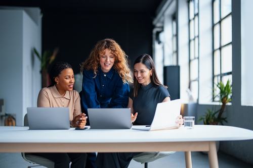 Three women collaborate around laptops at a modern office table, smiling and engaged. One stands leaning in while two are seated. Large windows and plants brighten the contemporary workspace.