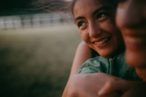 A young woman in a teal top smiles warmly while embracing someone in the foreground. Her hair blows slightly in the wind, with a blurred green field and fence visible in the soft background.
