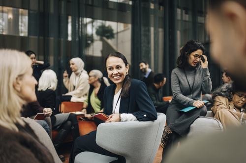 A smiling woman in a navy blazer sits among a diverse group of professionals at an indoor conference or seminar, holding a notebook and engaging with those around her in a modern, glass-walled venue.
