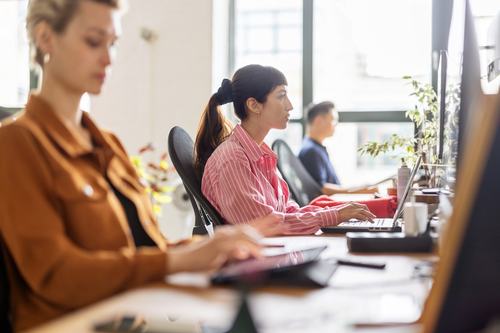 Three office workers sit in a row at a long desk, focused on their computers. A woman in a pink striped shirt types on a laptop, flanked by a colleague in an orange jacket and another in a blue shirt near bright windows.