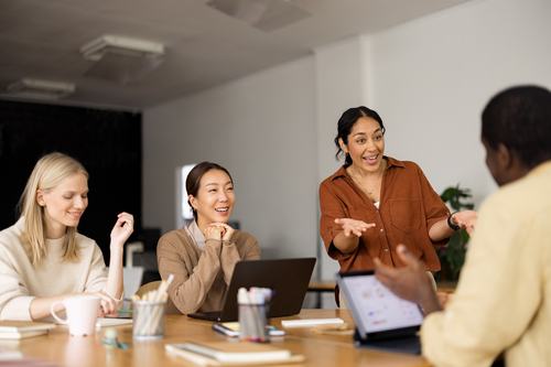 Four diverse women collaborate in a bright office meeting. One stands gesturing enthusiastically while others listen and smile. A laptop, tablet, and supplies are on the table, creating an engaging, positive team discussion atmosphere.