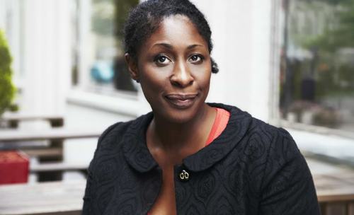 A woman wearing a black textured jacket over a red top, with short natural hair and small earrings, poses confidently in an outdoor seating area with a soft-focus background.