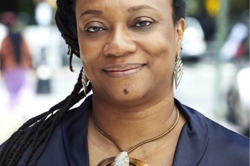 A woman with dreadlocks smiles warmly at the camera, wearing a navy blue top, gold hoop earrings, and a bold statement necklace featuring a large gold pendant with a crystal stone. Background is blurred outdoors.