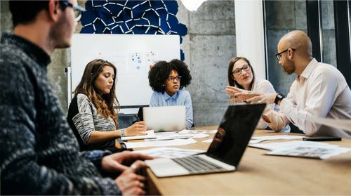 A diverse group of five professionals collaborating around a conference table with laptops and documents. A man gestures while speaking, others listen attentively. A whiteboard and bold blue wall art are visible in the background.