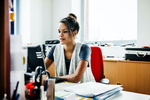A young woman with a bun hairstyle sits at an office desk, focused on her laptop. She wears a gray vest over a blue shirt, with papers and stationery nearby. A bright window is visible in the background.