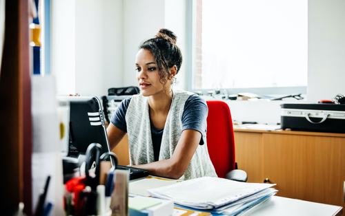 A young woman with a bun hairstyle sits at an office desk, focused on her laptop. She wears a gray vest over a blue shirt, with papers and stationery nearby. A bright window is visible in the background.