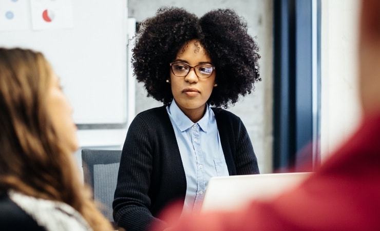 A woman with natural curly hair and glasses sits attentively in a meeting, wearing a light blue shirt and black cardigan, with a laptop open in front of her and colleagues visible in the foreground.