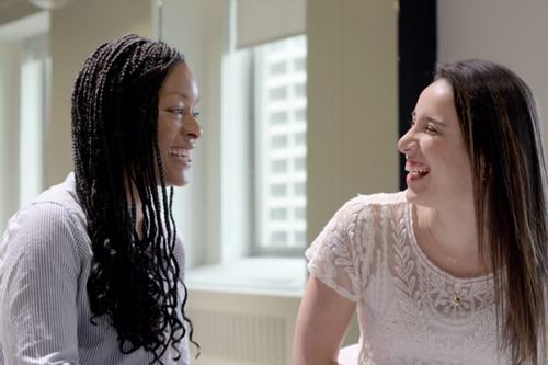 Two women smile and chat together in a bright office space, one holding an open magazine. One wears a striped shirt with braided hair; the other wears a white lace top with straight dark hair.