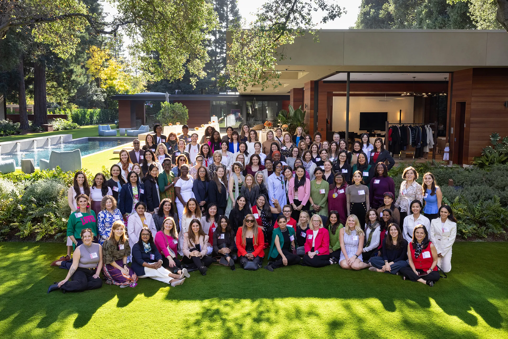 A large group of women pose for a photo on a lawn at an upscale outdoor venue with a pool and modern building. Many wear name badges, suggesting a professional conference or networking event on a sunny day.