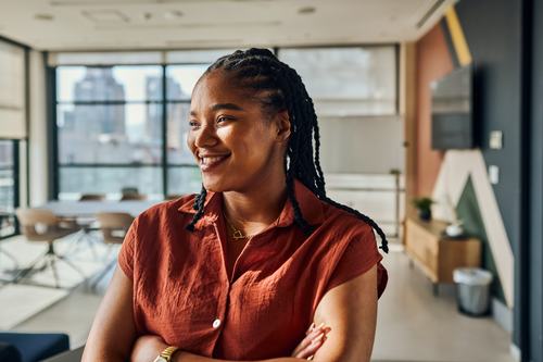 A confident young woman with braided hair smiles warmly in a modern office, arms crossed, wearing a rust-red shirt and gold jewelry, with a city skyline visible through large windows behind her.