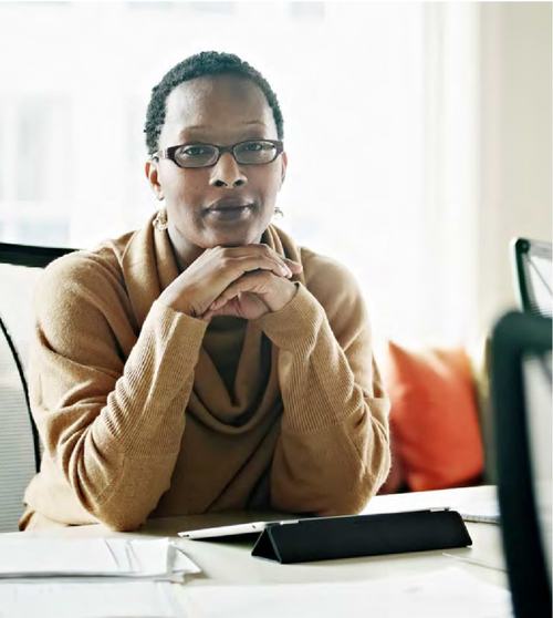 A professional woman wearing glasses and a tan sweater sits at a desk with her hands clasped, looking confidently at the camera. A tablet and papers are on the desk behind her.
