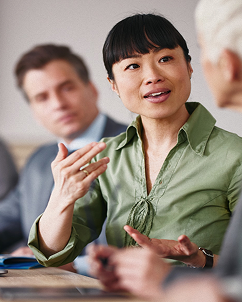 An Asian woman in a green blouse speaks animatedly during a meeting, gesturing with her hands. A man in a suit is visible, blurred in the background.