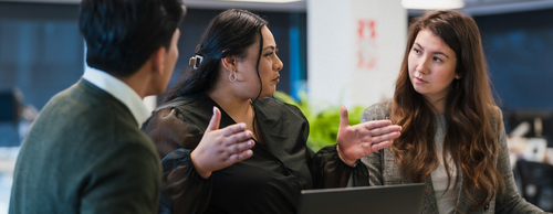 Three colleagues engaged in a discussion at a modern office, with a woman in a black top gesturing expressively while speaking to a man and another woman in a blazer attentively listening.